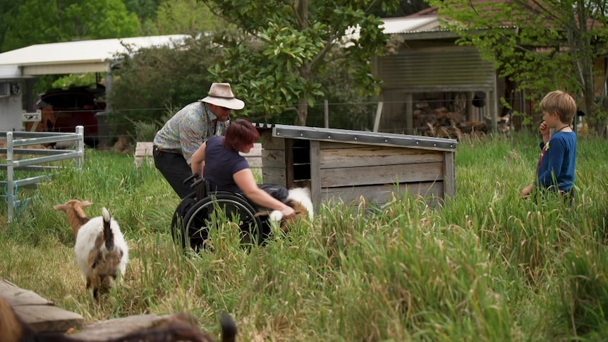 Image of a woman holding a goat while in a wheelchair with a man and child in the foreground.