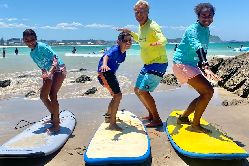 A surfing instructor shows three kids how to stand up on a surfboard.