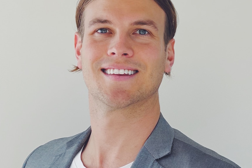 A head and shoulders image of a young man in a white t-shirt and grey blazer.