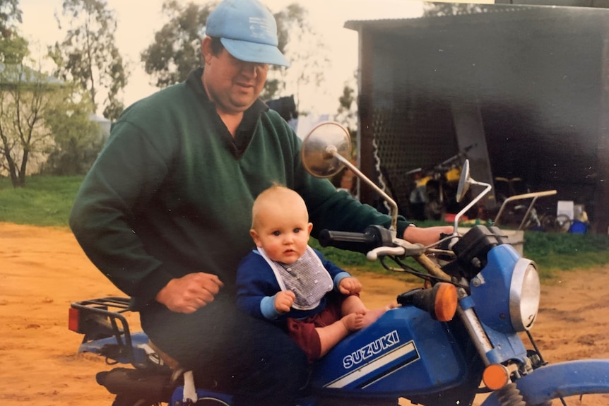 A man sits on a blue motorbike with his baby son sat in front of him. The baby looks to the camera, the dad looks down at son 