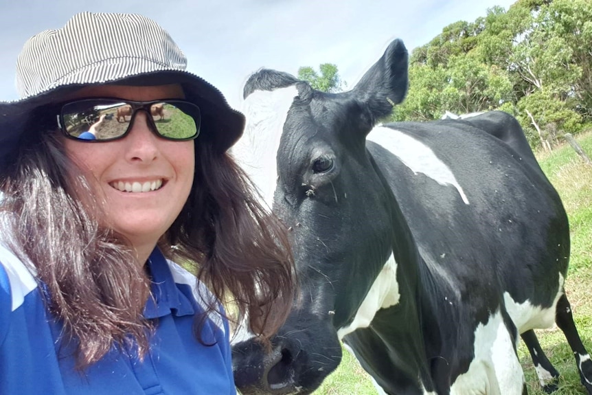 A smiling woman in a floppy hat, reflective sunglasses, stands in a field, next to a black and white cow who nuzzles her.