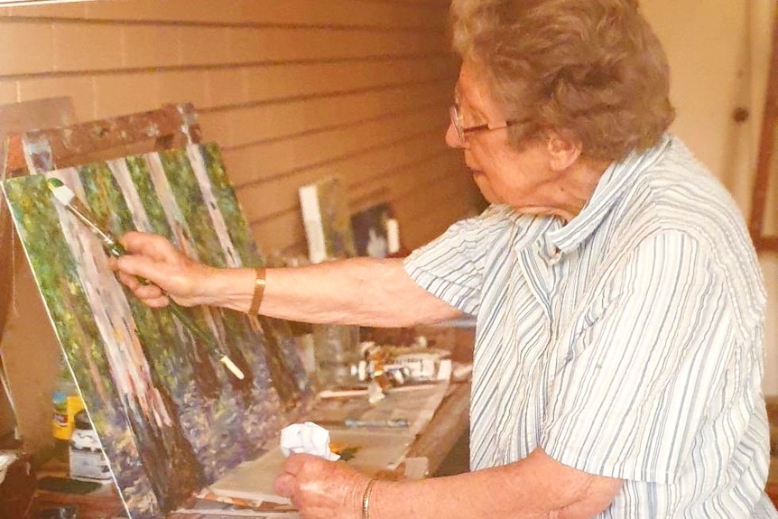 Older lady on the right painting a picture of tree trunks on the left.