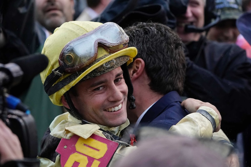 A jockey smiles at the cameras while hugging connections after his horse is named winner of a race.