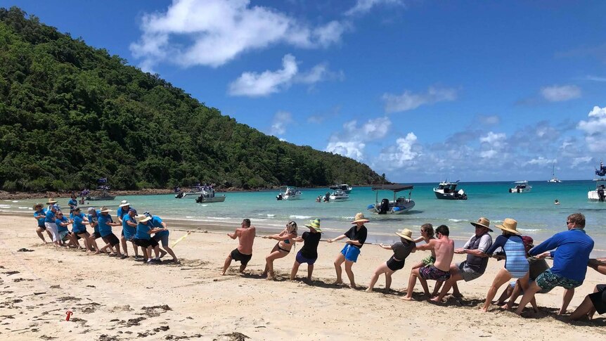 people play tug of war on a white sandy beach with boats in a bay  beyond them