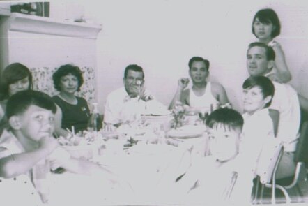 A black and white photo shows several children, women and men sitting around a table