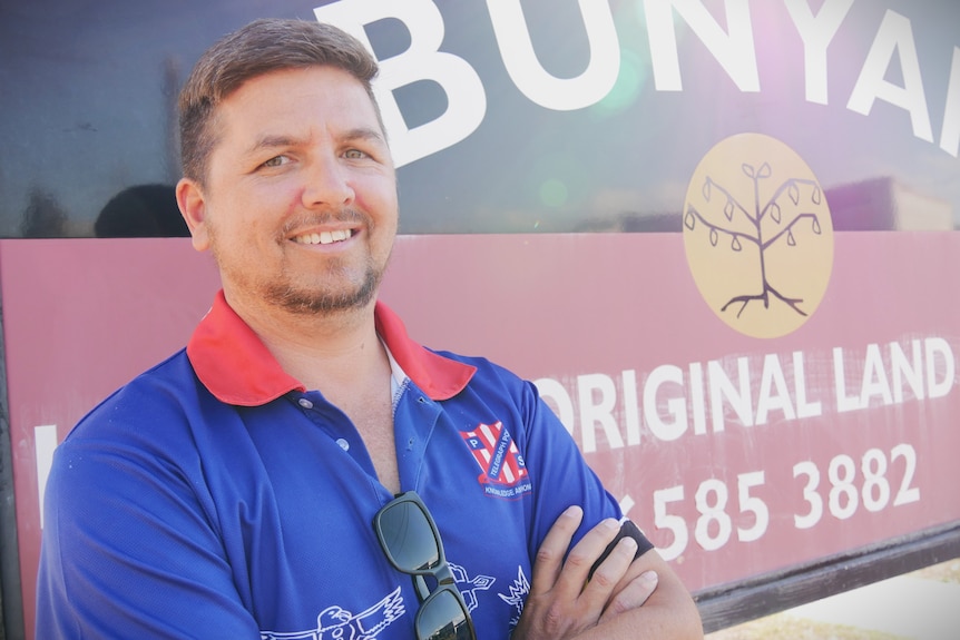 Young man with blue and red shirt looking and smiling at camera, with an aboriginal flag in background