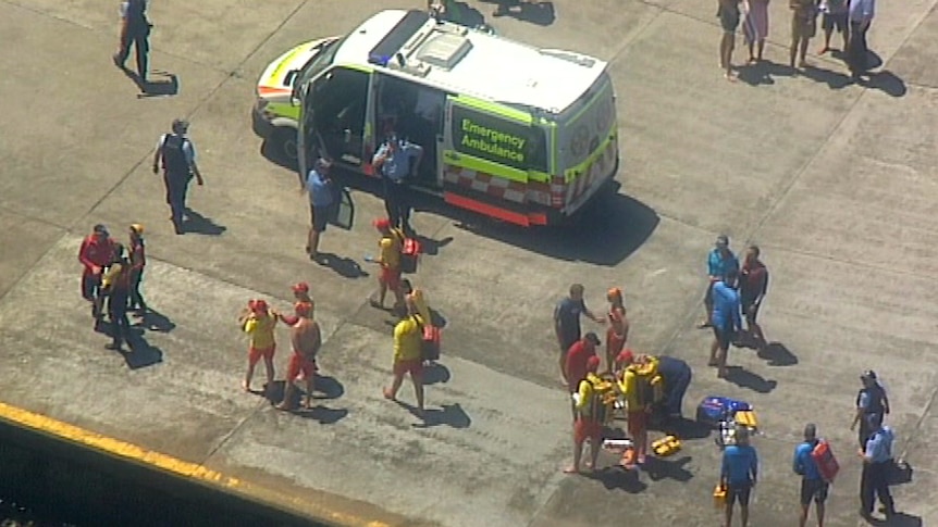 Lifeguards and lifesavers stand nearby an ambulance vehicle as police officers monitor the scene.