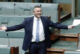 Darren Chester, wearing a dark suit, stands up in Parliament with his arms outstretched and a big grin on his face.