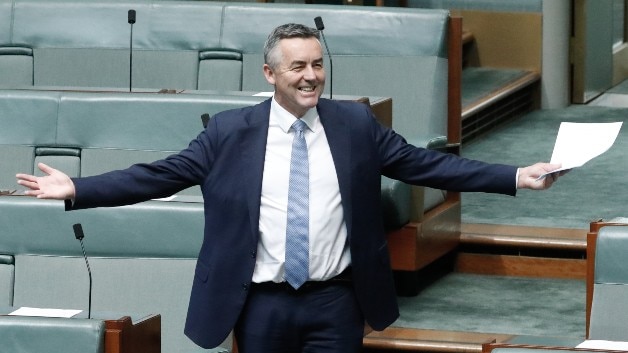 Darren Chester, wearing a dark suit, stands up in Parliament with his arms outstretched and a big grin on his face.