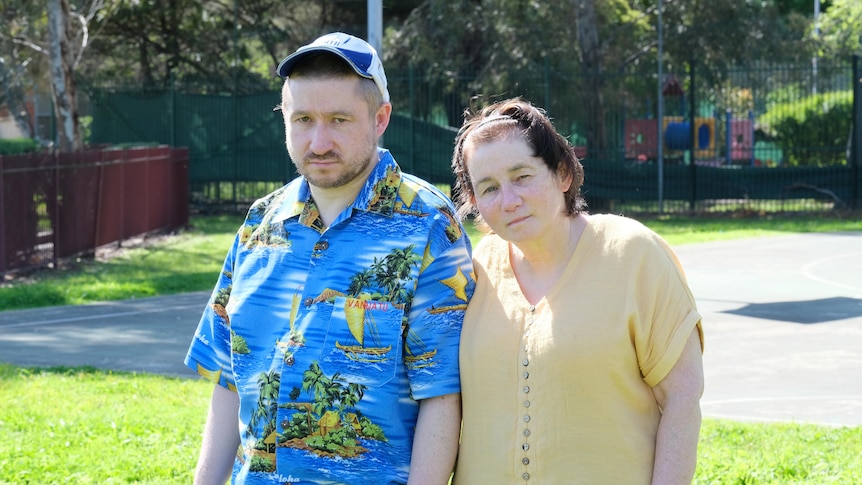 A young man with light facial hair wears a blue cap and Hawaiian shirt, stands with older woman in a light top.