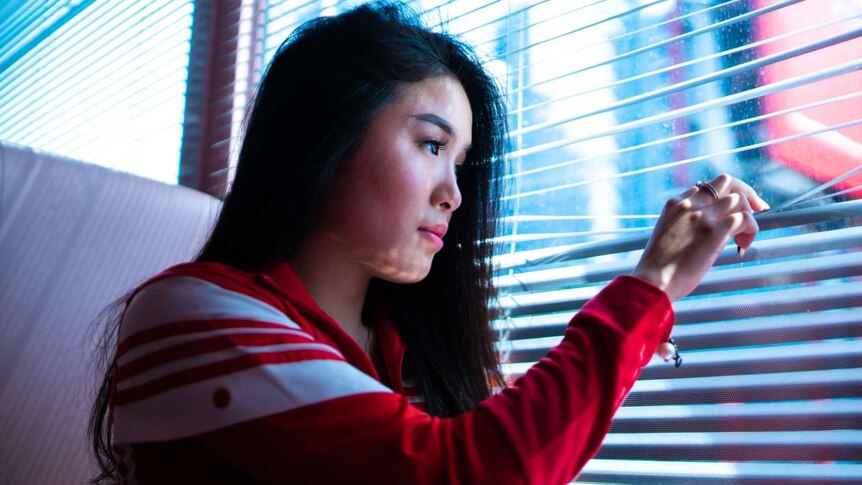 A young woman is looking through vertical blind on a window, like she's a bit anxious to go outside.
