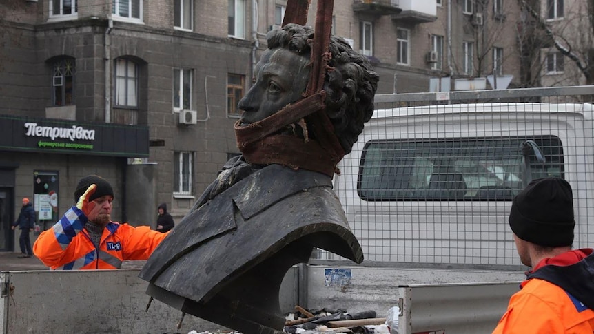 Two men in bright orange hi-vis clothing assist a large metal bust of Alexander Pushkin into the back of a ute.