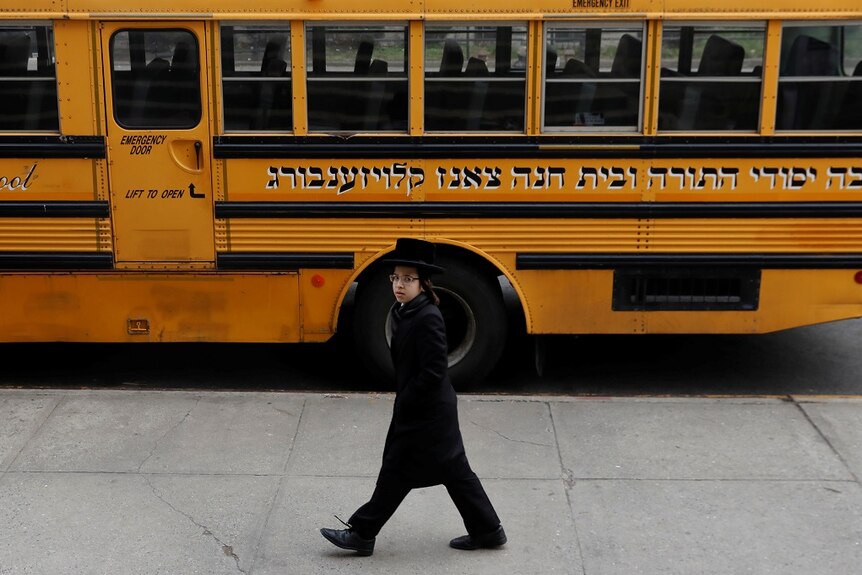 A young Jewish boy wearing a black hat, coat and slacks has his hands in his pockets as he walks down a footpath.