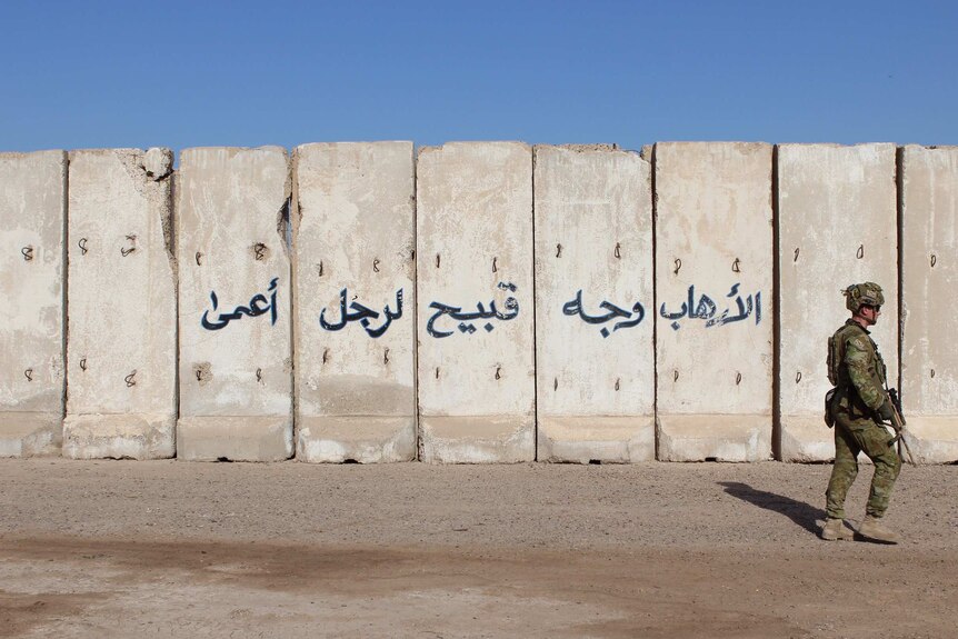 A soldier walks past a wall with Arabic writing on it.