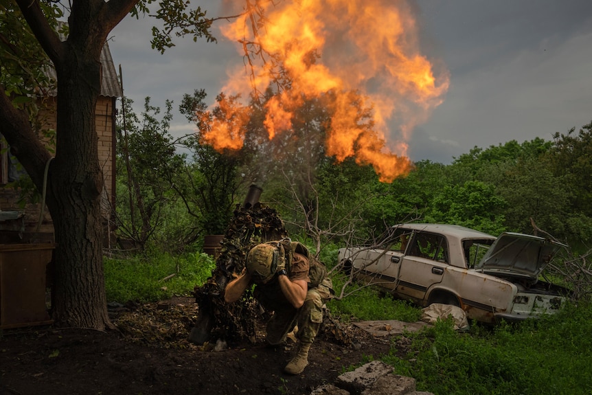 A Ukrainian soldier covers his ears as he fires a mortar at Russian positions next to a burnt out car.
