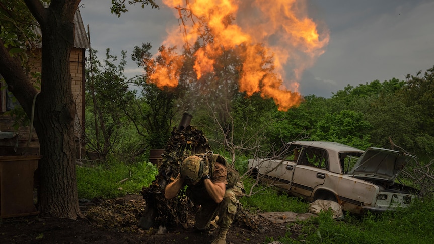 A Ukrainian soldier covers his ears as he fires a mortar at Russian positions next to a burnt out car.