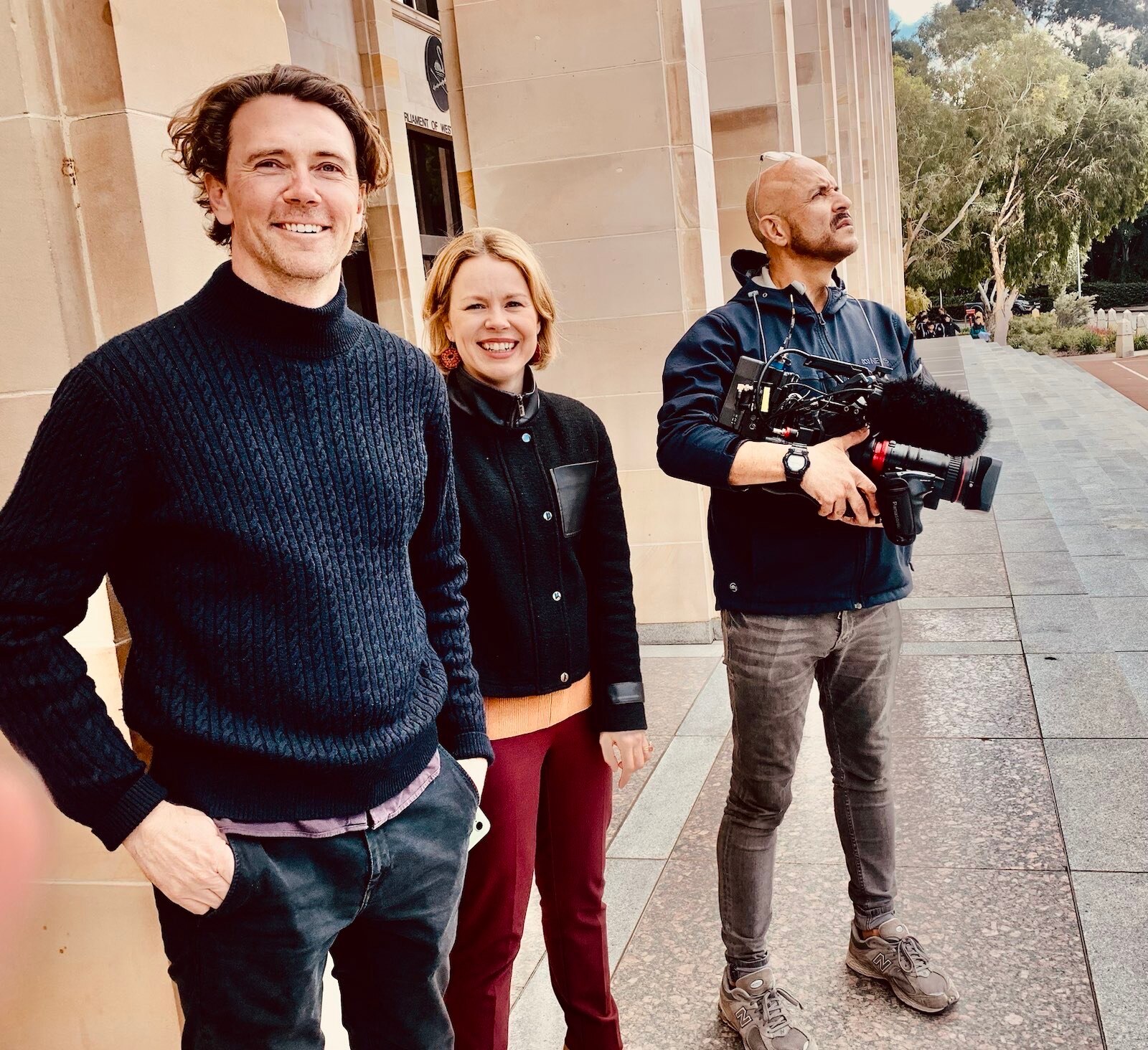 Woman standing between two men, one holding a camera outside a building.