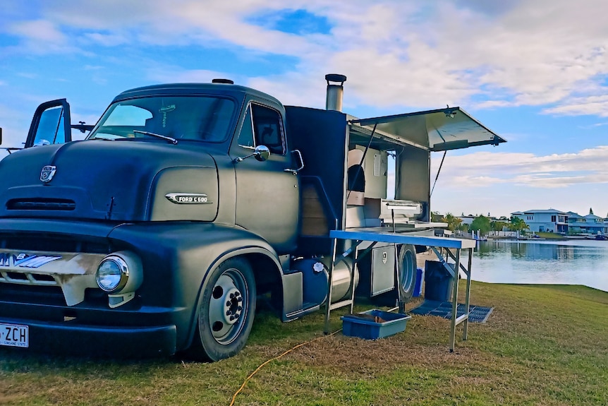 An old school, black Ford truck with a modified wood fire pizza oven on the back parked next to a lake