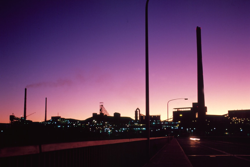 Dark buildings are silhouetted against a purple sunset with smokestacks from a mine visible.
