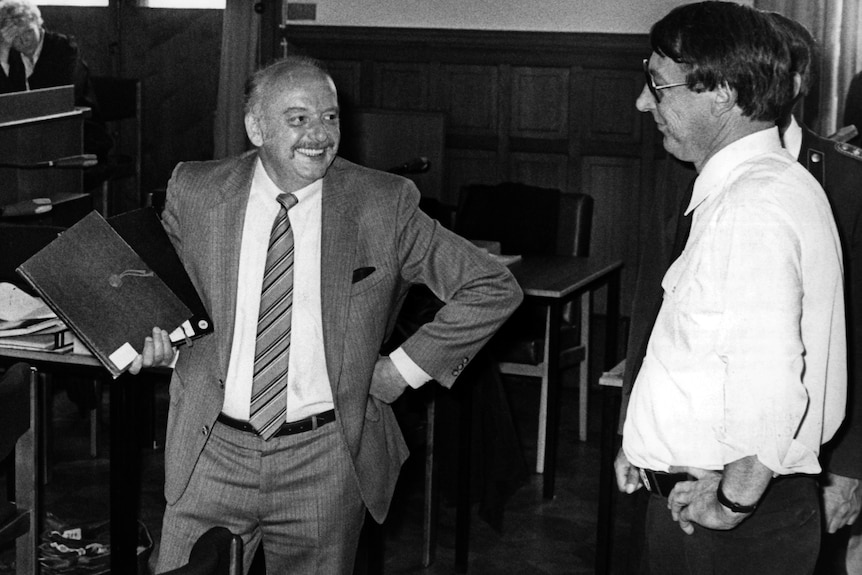 Black and white photo of Konrad Kujau, in suit and tie, smiling widely and holding a large book in one hand.