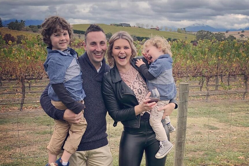 A man and his wife hold each of their children, little boys, outside at a vineyard and the whole family is smiling big