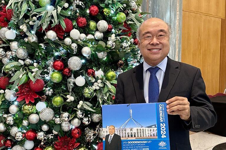 A man in a suit holds a blue calendar in front of a Christmas tree.
