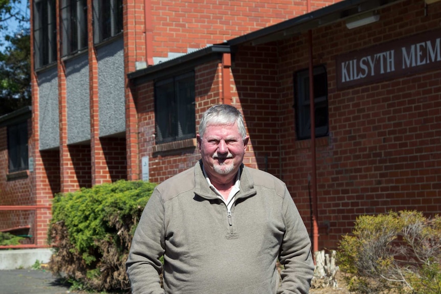 A man with grey hair and a white goatee stands in front of a large red brick building.