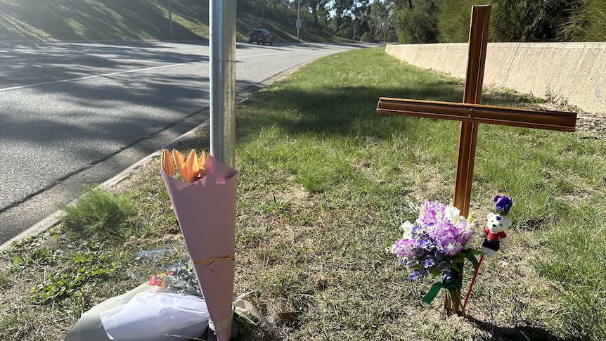 A grass strip next to a road with multiple bouquets of flowers and a wooden cross.