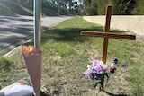 A grass strip next to a road with multiple bouquets of flowers and a wooden cross.