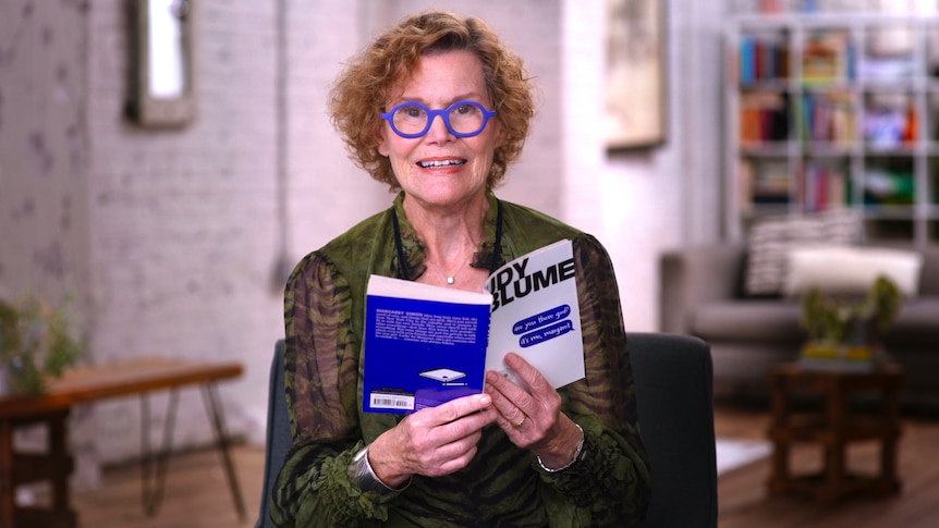 The author Judy Blume - a woman in her mid 80s with curly red hair and blue glasses - sits and reads from a book