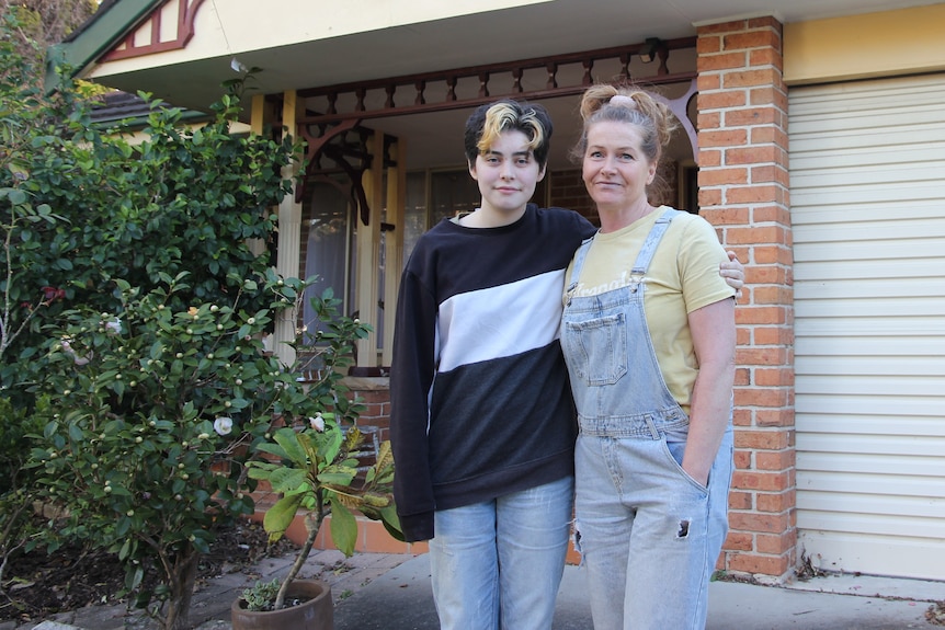 Sage and Bec Nikolovski stand arm in arm outside their home, smiling at the camera.