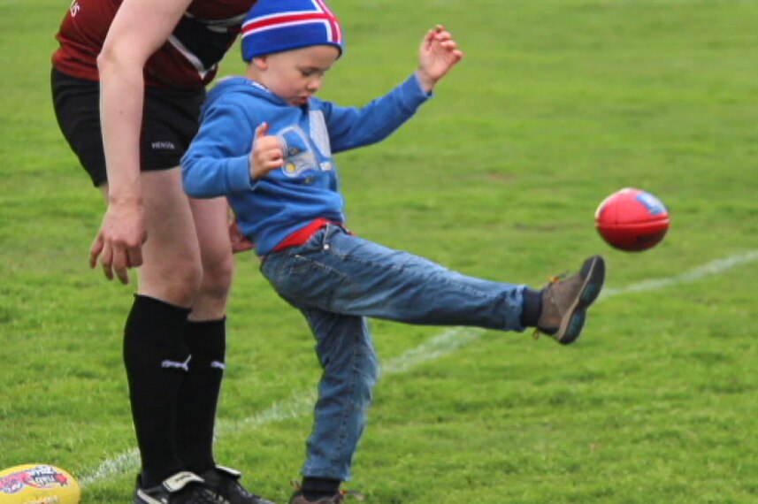 A boy wearing jeans, a blue jumper and an Iceland beanie kicks a red Aussie Rules football with a man standing behind him.