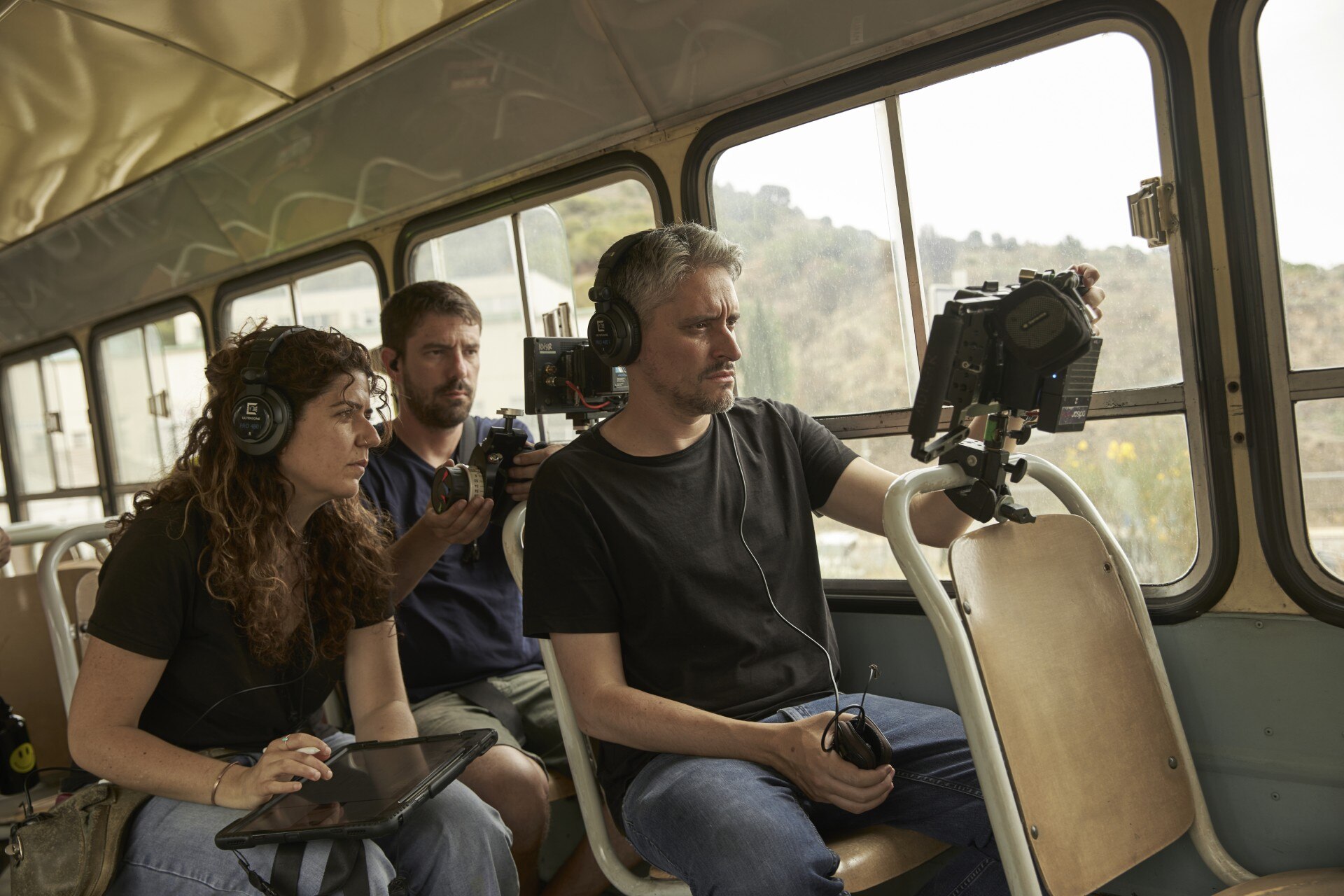 A film crew wearing headphones and looking at monitors sit in a bus.