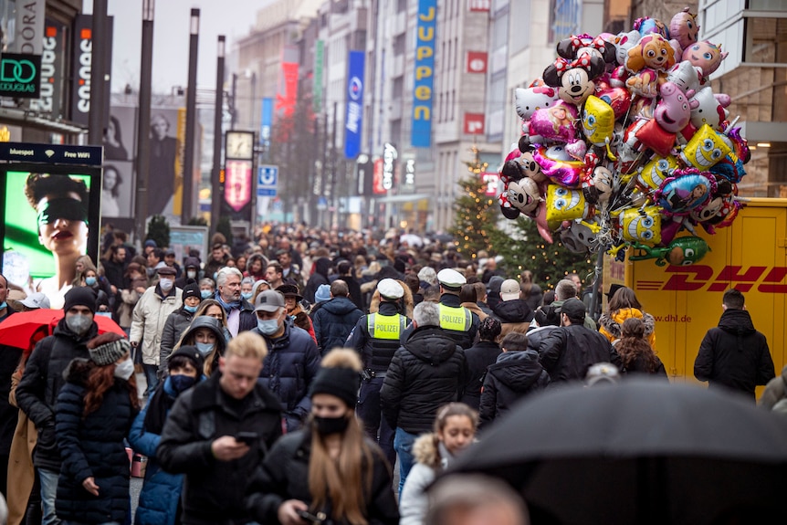 Hundreds of people wearing masks walking on a busy city street with Christmas trees in the distance