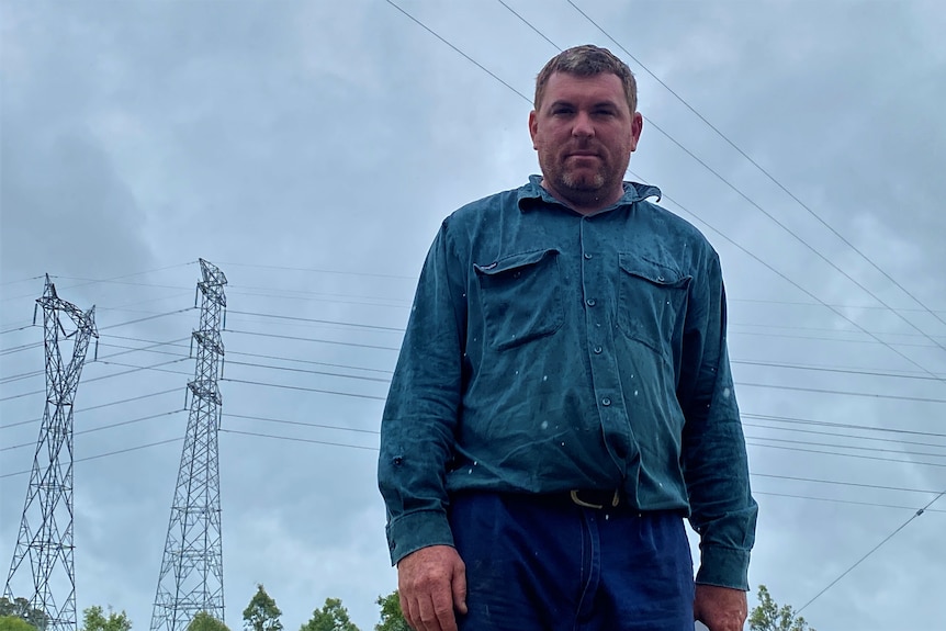 A man stands seriously with transmission towers and electricity cables towering over him. The sky is grey with storm clouds 