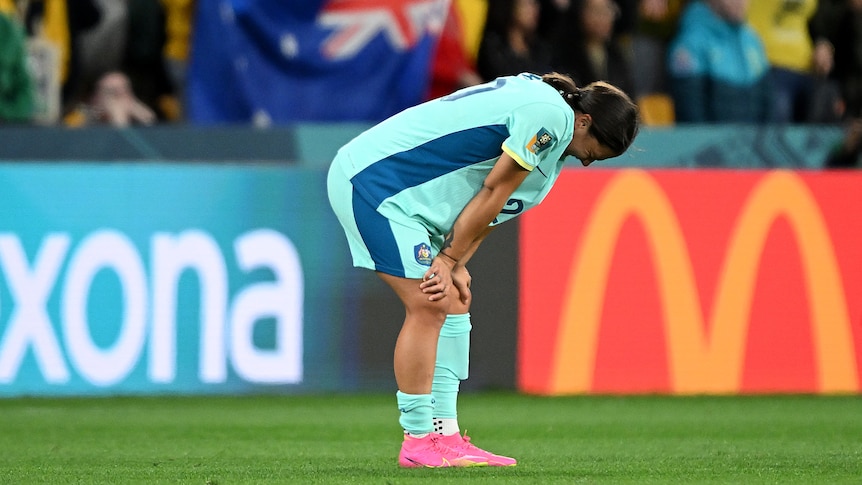 A soccer player wearing blue and light green leans on her knees with an Australian flag in the background