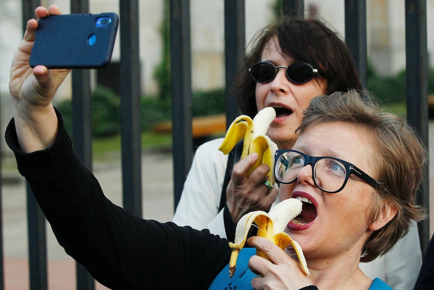 A woman with short hair wearing black-rimmed glasses and red lipstick takes a selfie on her phone as she eats a banana.