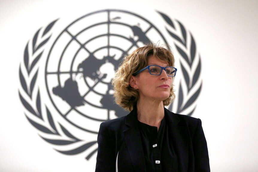 Agnes Callamard wears a black blazer and shirt while standing in front of a large black UN logo on a white wall.