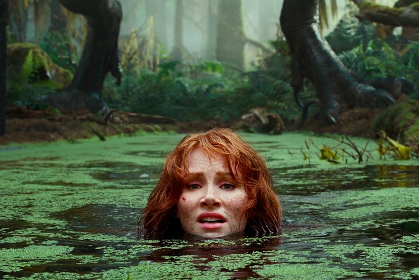 White woman with fiery red hair looks distressed, submerged in moss-topped pond. Large dinosaur feet can be seen on bank beyond.