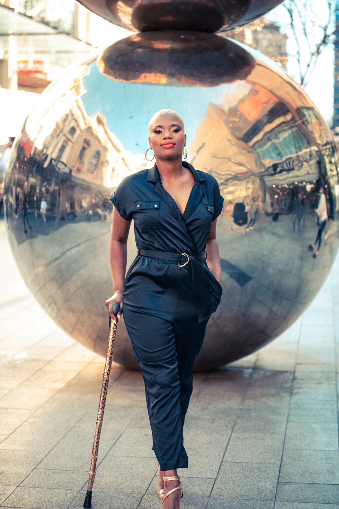 Khadija stands in front of large reflective metallic balls. She has a glittery walking stick and proud look on her face.