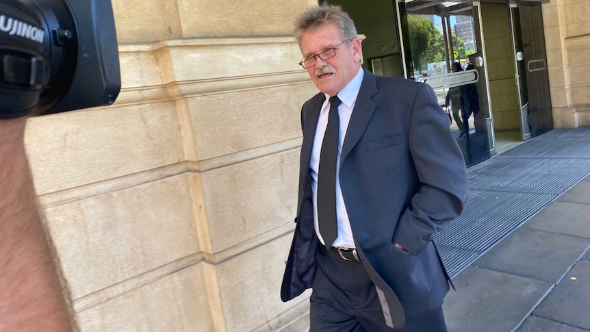 A man in a black suit wearing a white shirt and glasses walks past a television camera