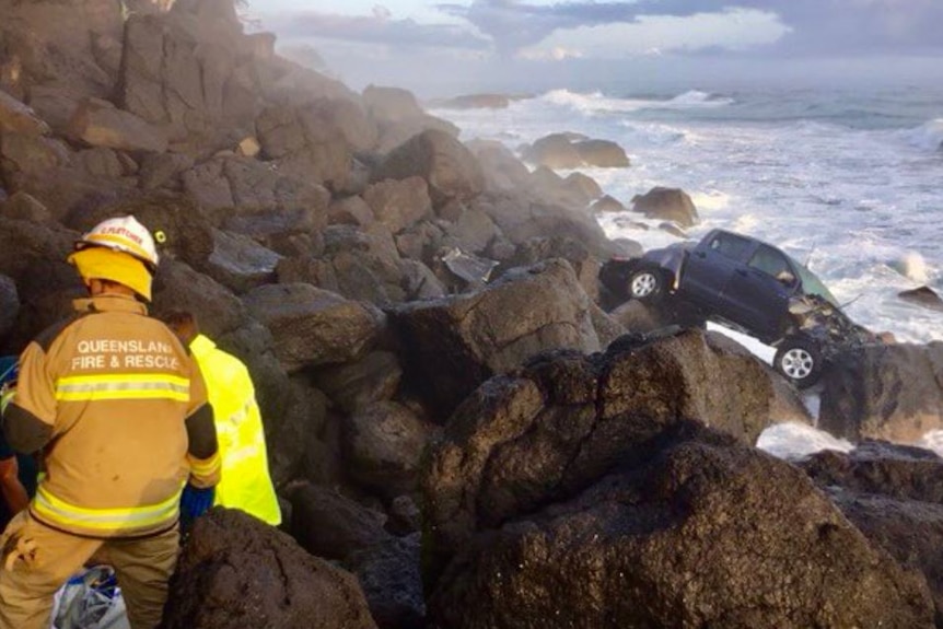 A smashed ute is close to the ocean and choppy waves at the bottom of rocks