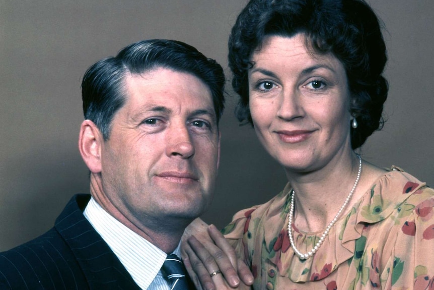 Paul Cronin as Dave Sullivan sits while Lorraine Bayly as Grace Sullivan stands beside him, resting her hands on his shoulder.