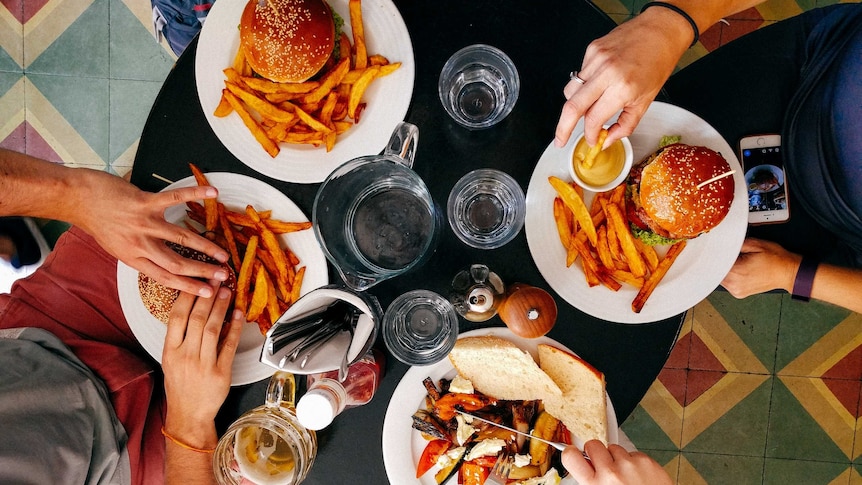 View from above of hands reaching out to eat from a table covered in plates of burgers and fries.