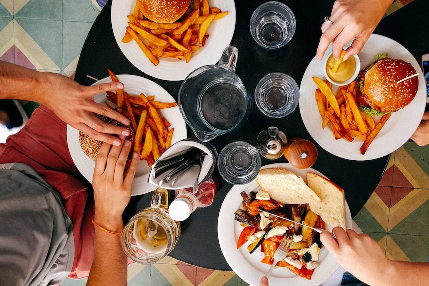 View from above of hands reaching out to eat from a table covered in plates of burgers and fries.