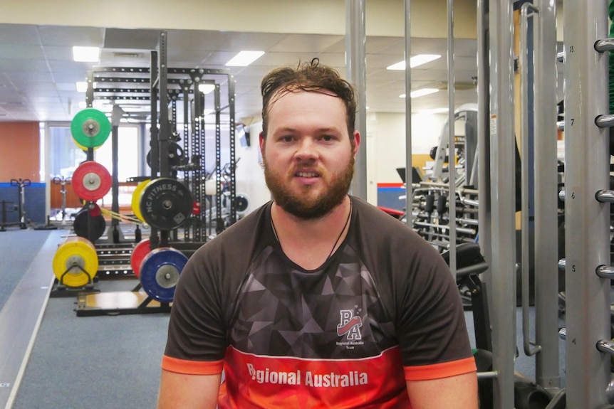 A man sits in a gym, weights can be seen in the background. He is smiling at the camera. 