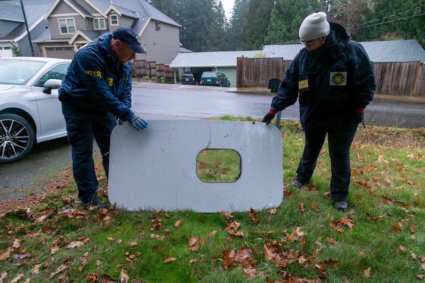 A medium shot of two people holding up a door plug from an airplane on the front lawn of a house after it fell to Earth.