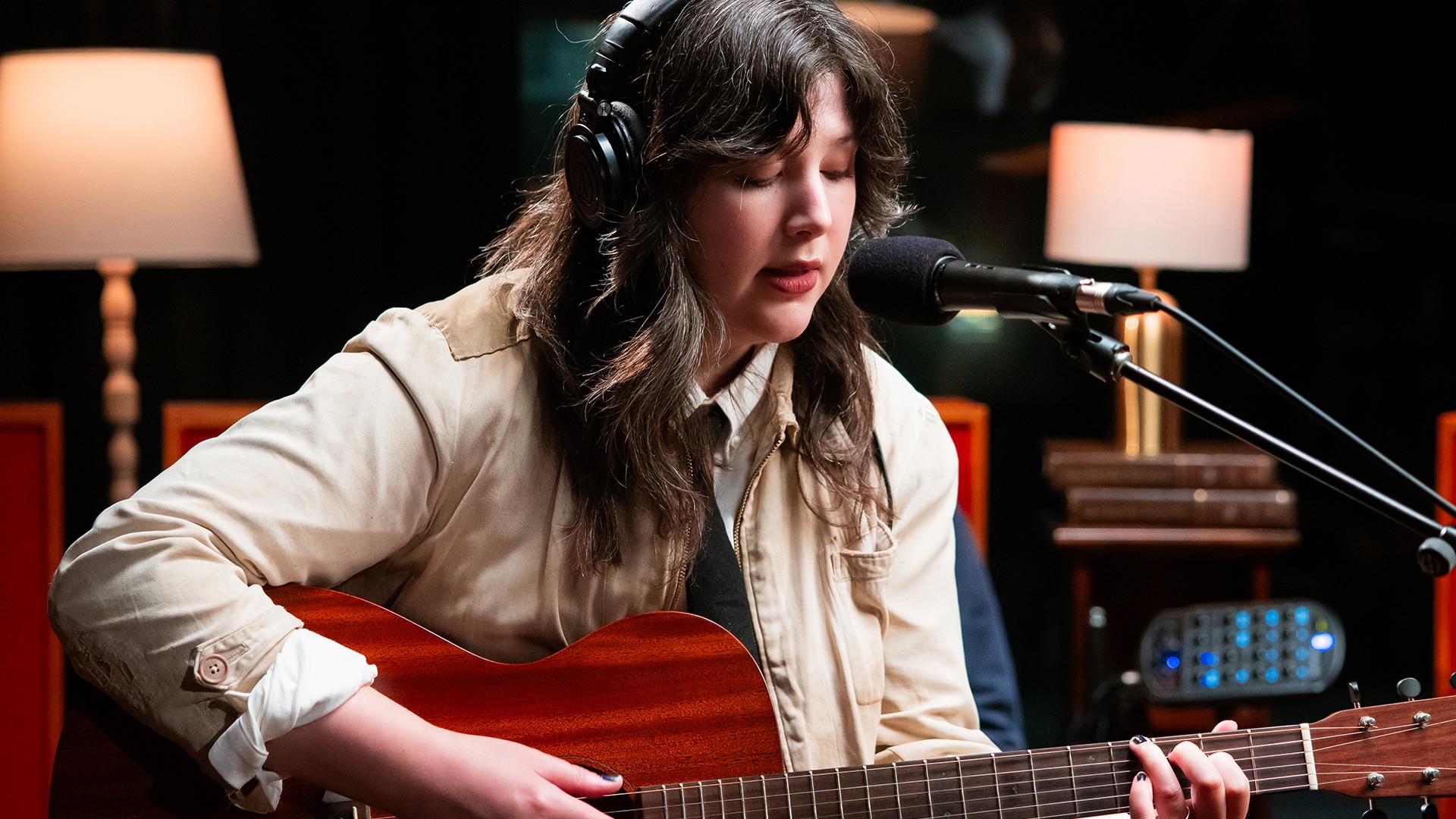Lucy Dacus plays an acoustic guitar in a studio. She's wearing headphones and sings into a microphone