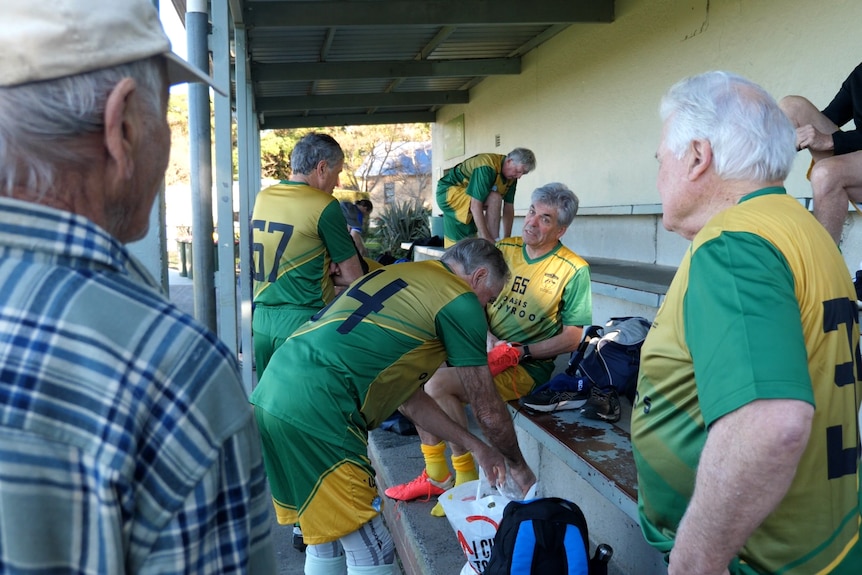 a team of older men in football jerseys sit on a bench talking to each other