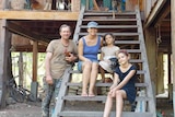 Ben and Sharyn Davis sit with their two daughers, a dog, and chicken on stairs outside their wooden home.